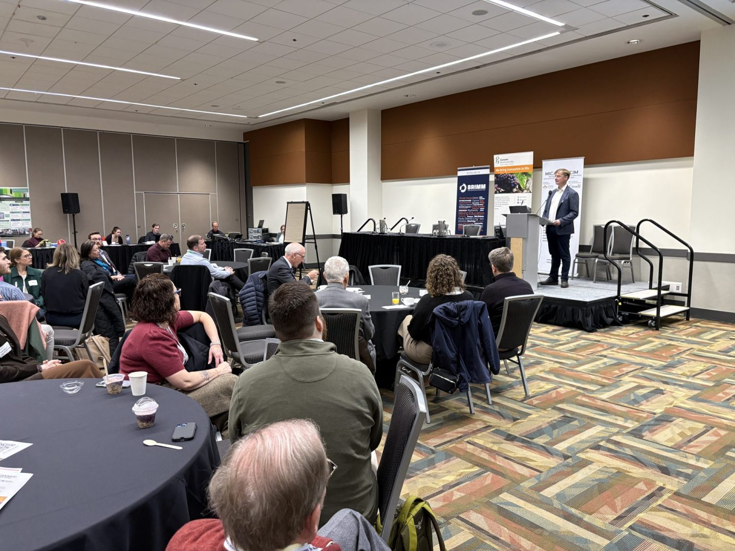 A speaker at a podium addresses an audience of people seated at round tables.